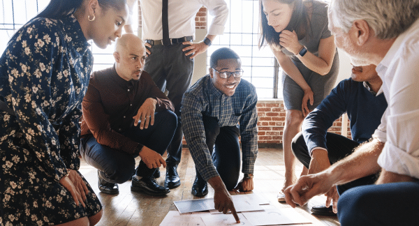 A diverse group of six professionals collaborates around a table, examining documents and discussing strategies in a bright, modern workspace.